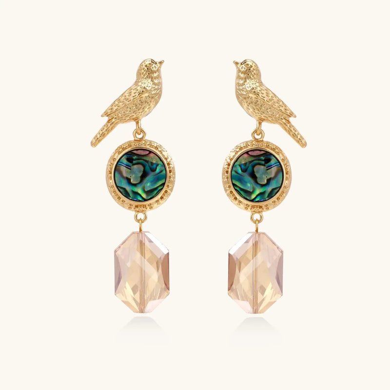 Gold-plated bird stud earrings with iridescent abalone shell medallions and champagne crystal drops on cream background